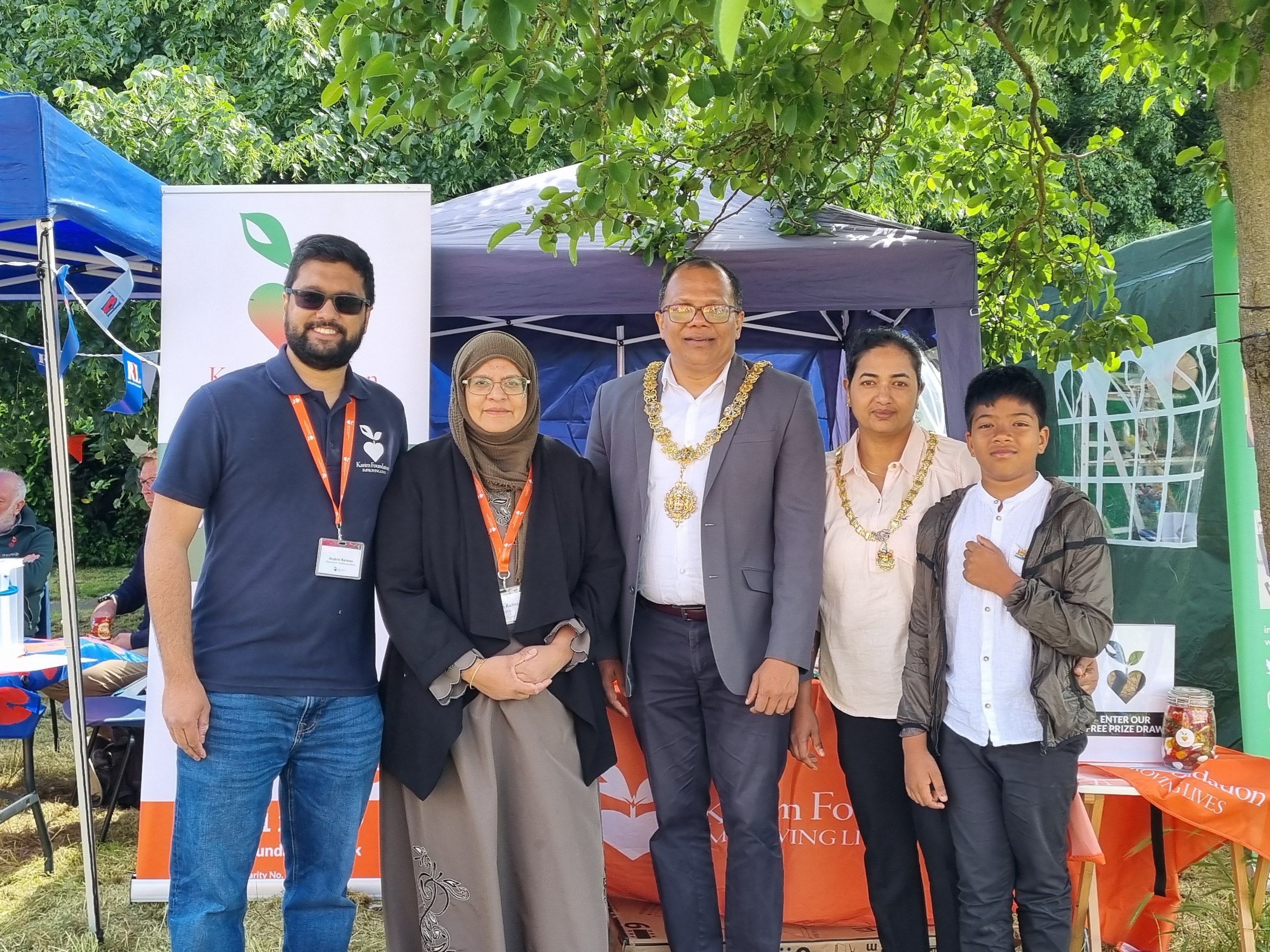 Ibrahim Rahman and Shahida Rahman on the left posing in front of the Karim Foundation stall with Councillor Baiju Thittala, the mayor of Cambridge, who is wearing a formal chain of office, typically worn by civic dignitaries. Standing to the mayor's left are his wife and son.